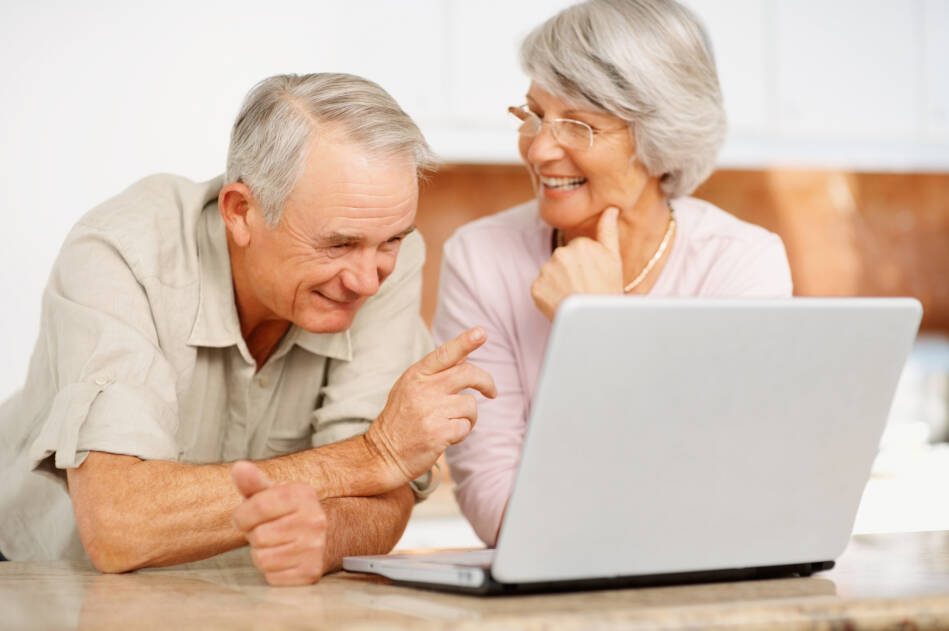 Una pareja de adultos mayores conversa sonriente frente a una computadora portátil